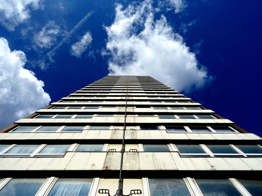 A tall multi-storey residential building seen from the ground with a clear blue sky and scattered white clouds above. The building's façade features multiple rows of windows, some with open blinds, and panels of weathered concrete or metal cladding. The perspective emphasizes the building's height, and there are no visible vans or moving equipment in the image. This image could relate to house removals and furniture transport, highlighting the challenges of moving large items within apartment buildings such as those by Man and a Van Bow, especially in no lift situations, although no moving activity is visible in this specific shot.