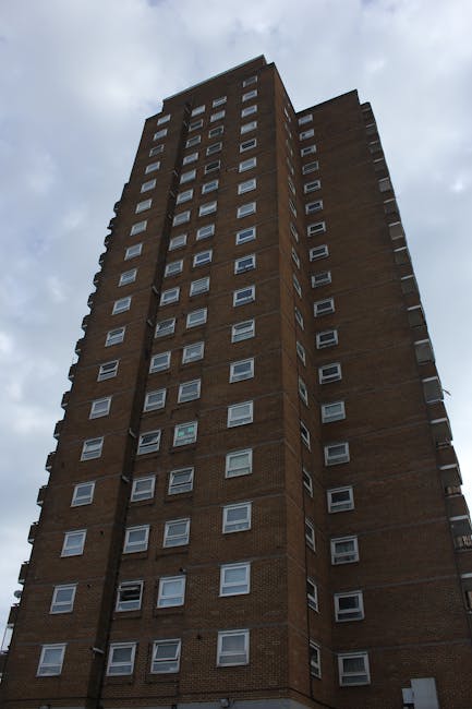 A tall multi-storey residential building with a brick façade and numerous white-framed windows seen from a low-angle perspective against a cloudy sky. The building features varied window arrangements, with some sections slightly recessed or protruding, indicating different architectural elements. The scene captures the exterior of the property before or during a home relocation, illustrating the typical high-rise environment for apartment moves. Man and a Van Bow, a professional removals company, commonly assist with such logistics in this area, offering packing, furniture transport, and loading services. The image emphasizes the height of the building, relevant to the challenges of loading and transporting belongings from upper floors during moving processes, especially where no lift access is available, aligning with Bow removals guidance for Bow Road flats.