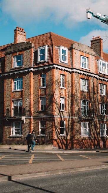 A tall multi-storey residential building seen from the ground with a clear blue sky and scattered white clouds above. The building's façade features multiple rows of windows, some with open blinds, and panels of weathered concrete or metal cladding. The perspective emphasizes the building's height, and there are no visible vans or moving equipment in the image. This image could relate to house removals and furniture transport, highlighting the challenges of moving large items within apartment buildings such as those by Man and a Van Bow, especially in no lift situations, although no moving activity is visible in this specific shot.
